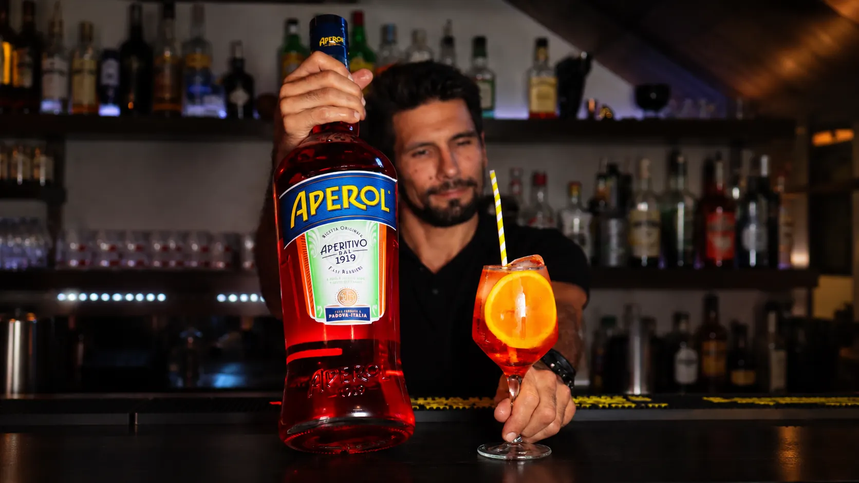 An Aperol Spritz with a slice of orange and a straw, beside a large bottle of Aperol, on the bar.