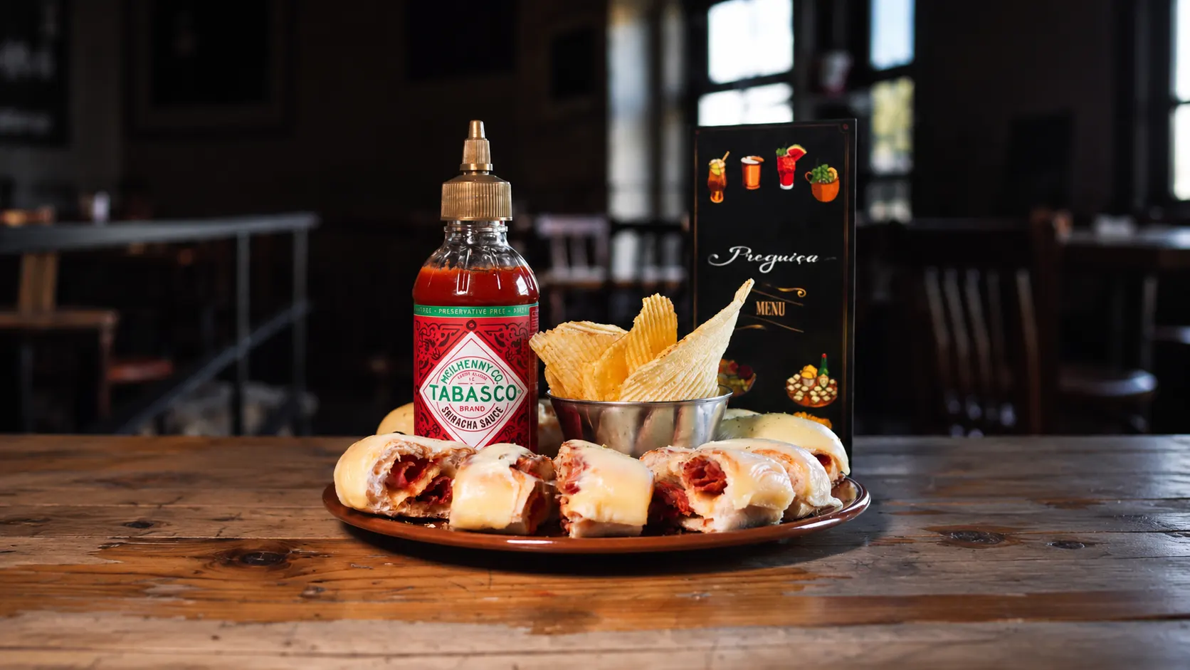 A platter of bread baked with chouriço and melted cheese, in pieces, with a bowl of crisps and a bottle of hot sauce.