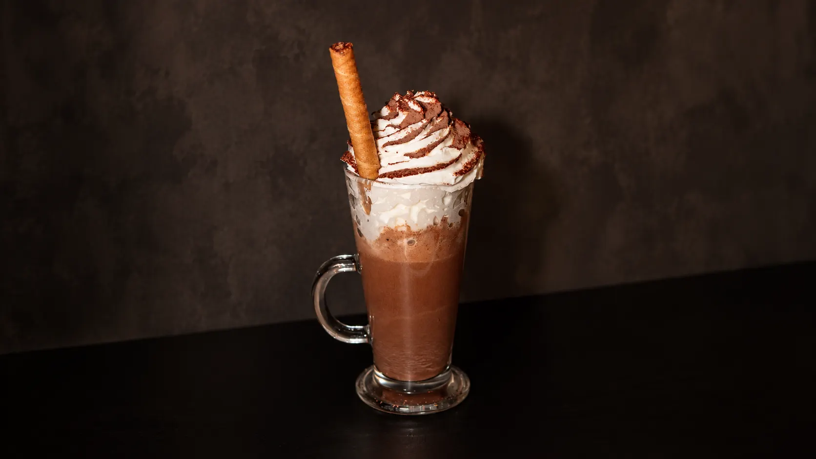 A hot chocolate in a glass mug, with whipped cream, cocoa and a wafer roll.