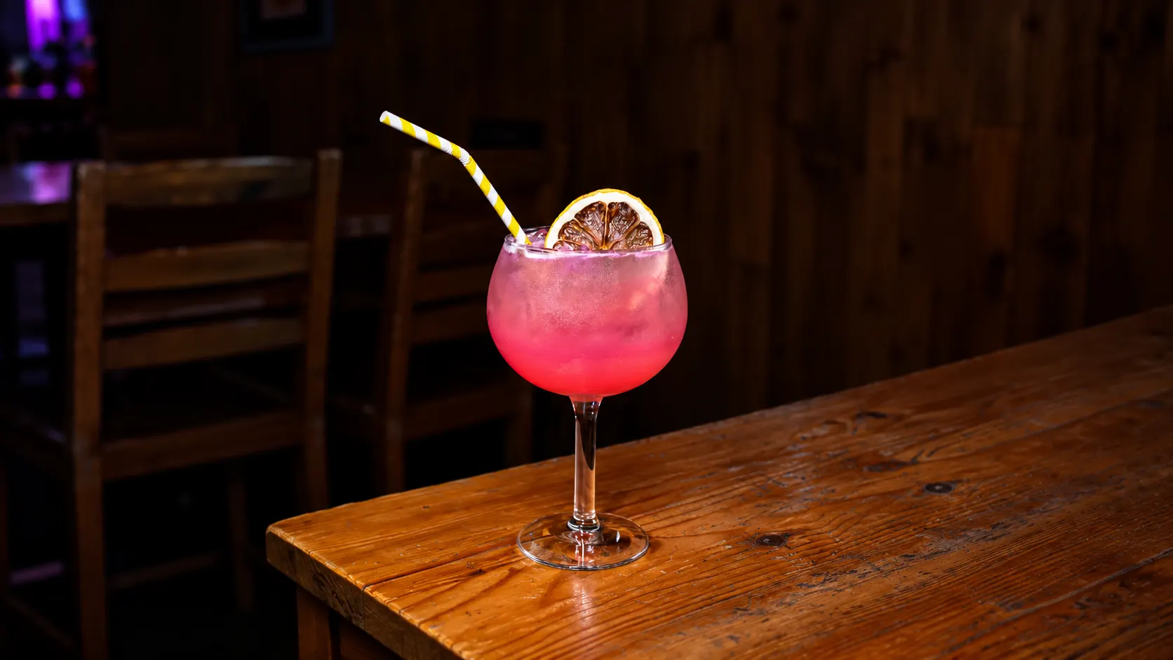 A balloon glass with a pink drink, a dried orange slice and a yellow-striped straw, on a wooden table.