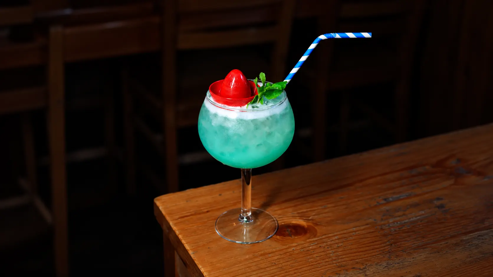 A balloon glass with a turquoise drink, crushed ice, a strawberry, mint and a blue-striped straw, on a wooden table.