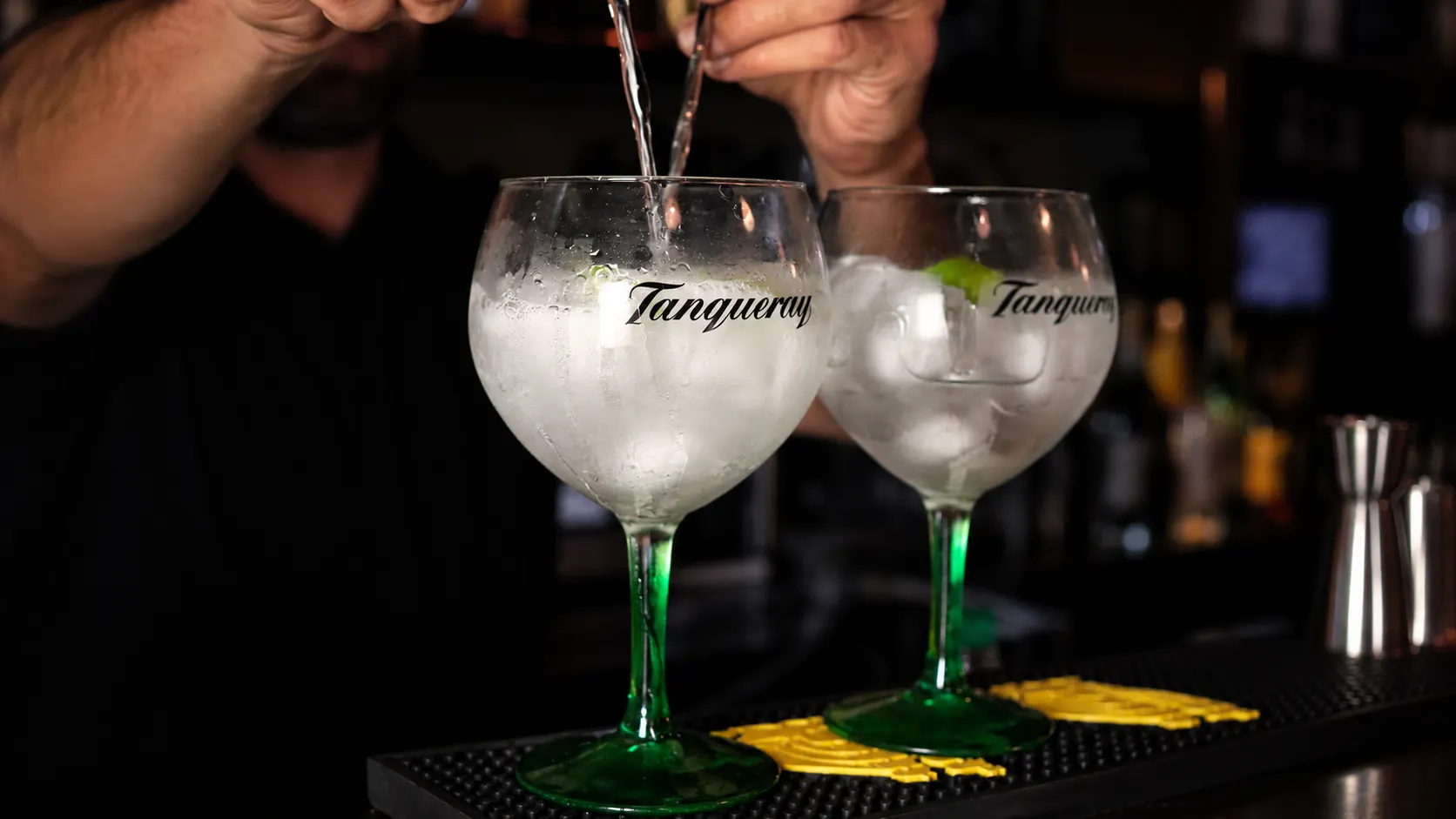 A gin and tonic being poured into balloon glasses engraved with the Tanqueray name.