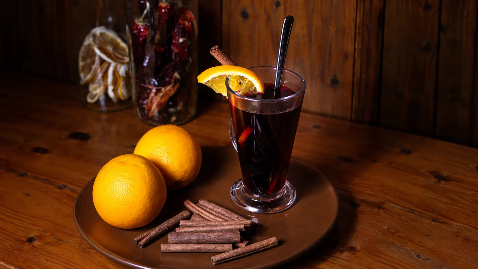 A Glühwein, hot red wine, in a glass mug with a slice of orange, beside oranges and cinnamon sticks.