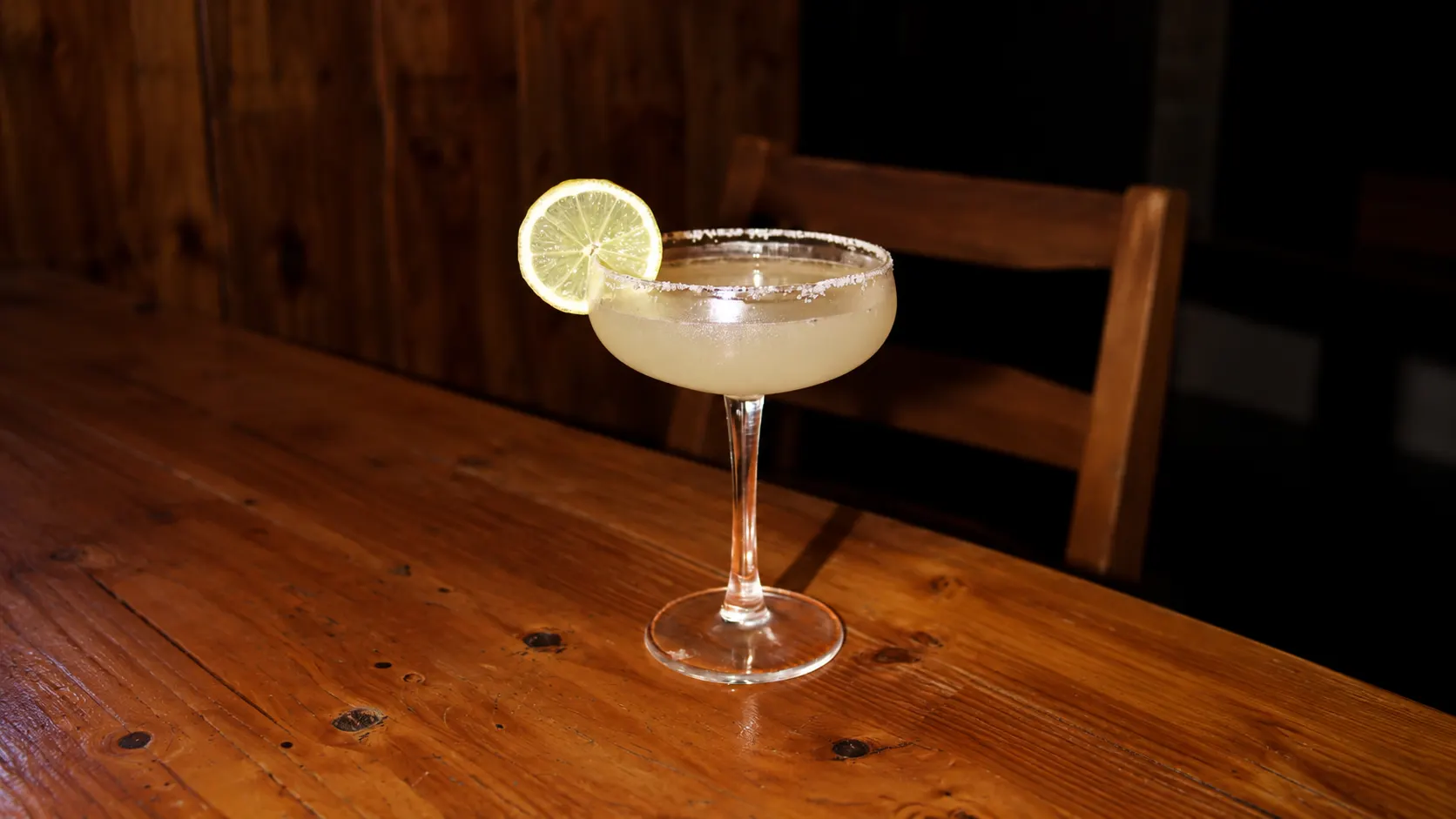 A margarita in a coupe with a salted rim and a slice of lime, on a wooden table.