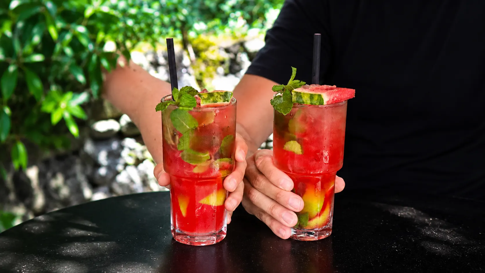 Two red watermelon mojitos with mint and a slice of watermelon, held in two hands.