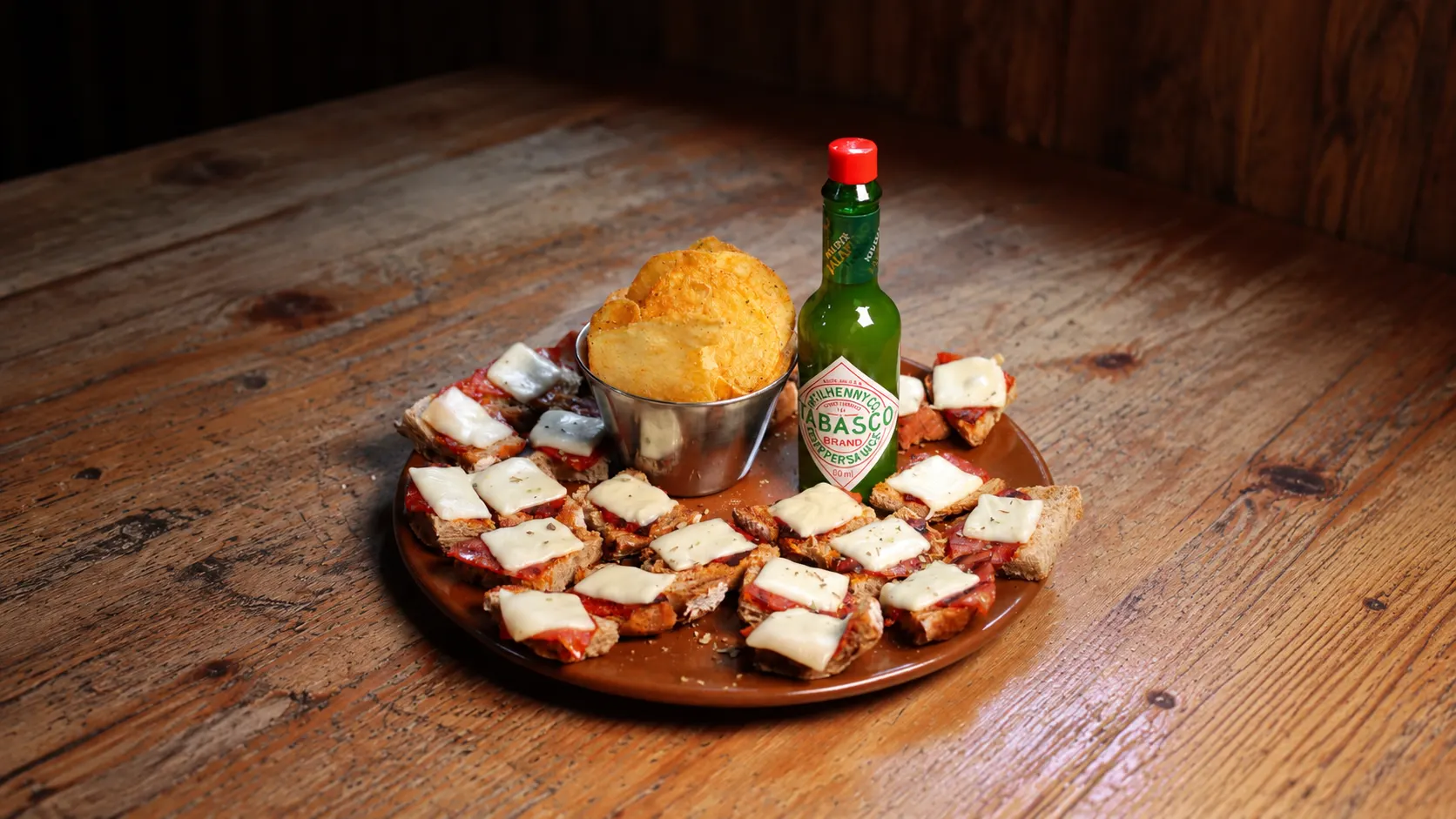 A platter of little toasts with chouriço and cheese around a bowl of crisps, with a bottle of Tabasco.