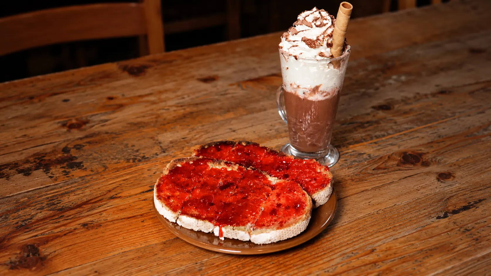 A hot chocolate with whipped cream and a wafer roll, behind two slices of toast with jam, on a wooden table.