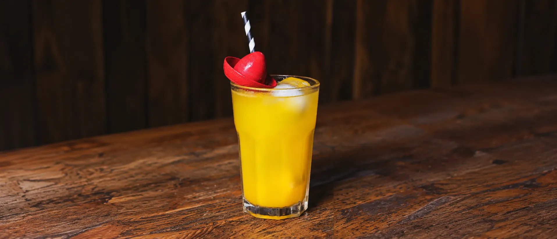 A yellow Unicorn in a glass tumbler with ice, a red decoration on the rim and a black-striped straw, on a wooden counter.