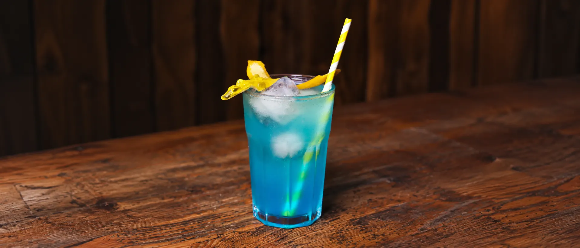 A blue Unicorn in a glass tumbler with ice, a yellow gummy shark, lemon peel and a yellow-striped straw, on a wooden counter.