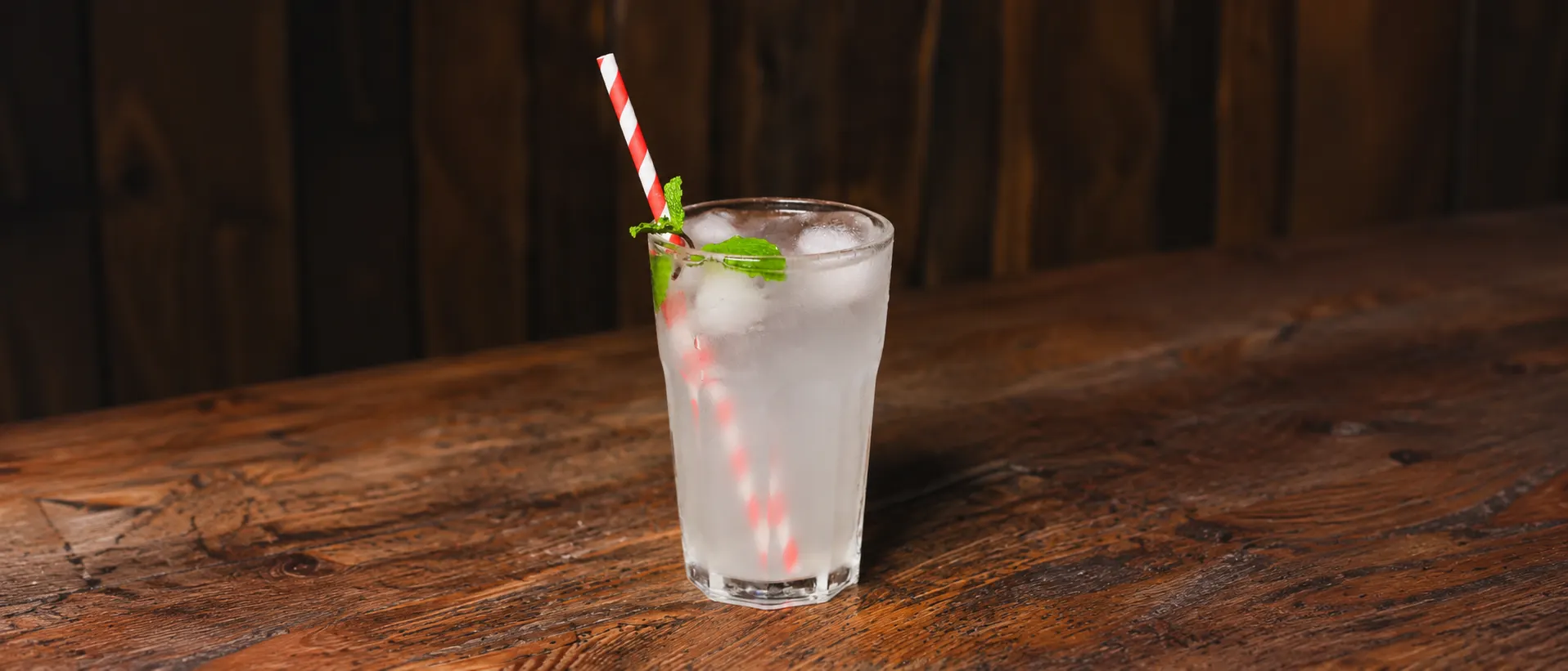A clear Unicorn in a glass tumbler with ice, a sprig of mint and a red-striped straw, on a wooden counter.