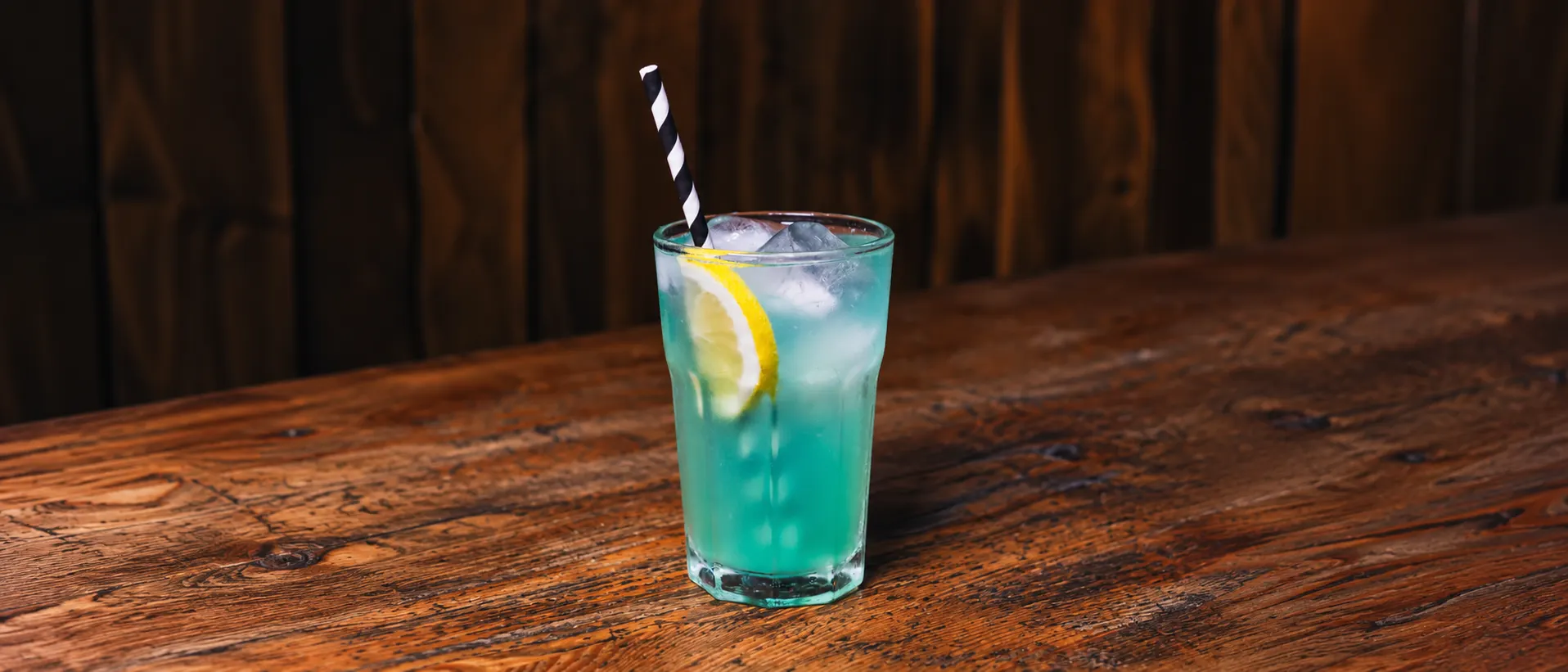 A turquoise Unicorn in a glass tumbler with ice, a slice of lemon and a black-striped straw, on a wooden counter.