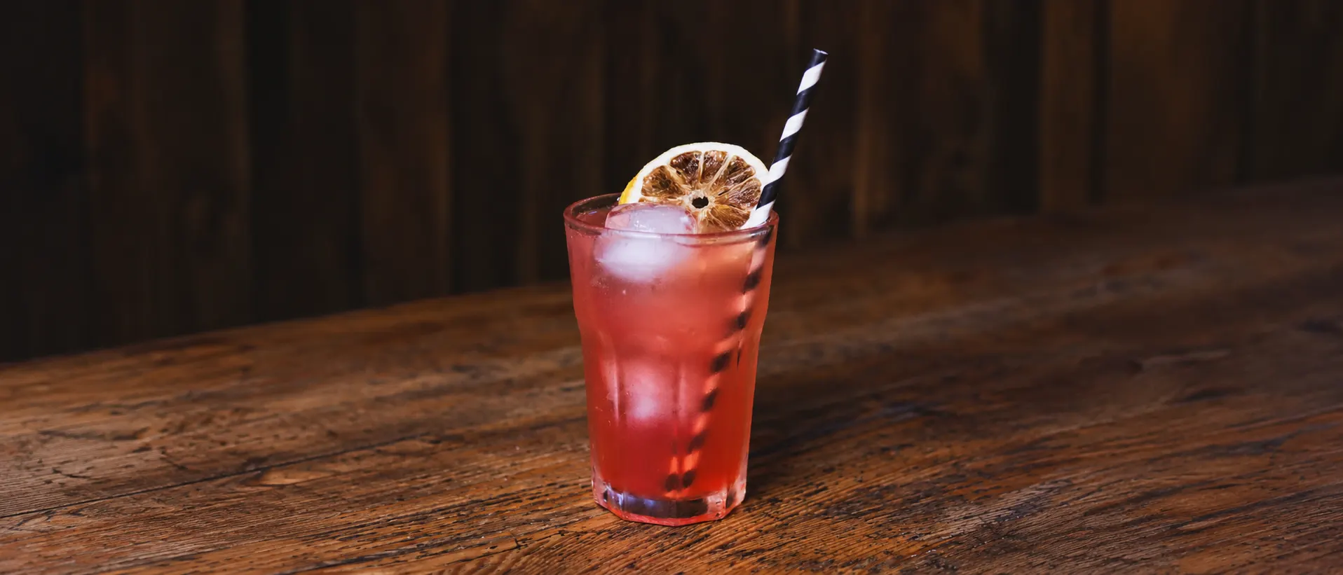 A red Unicorn in a glass tumbler with ice, a dried orange slice and a black-striped straw, on a wooden counter.