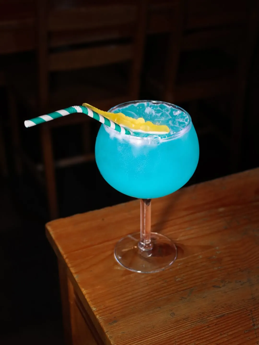 Um copo balão com uma bebida azul, gelo picado, uma fatia de fruta amarela e uma palhinha às riscas verdes, sobre uma mesa de madeira.