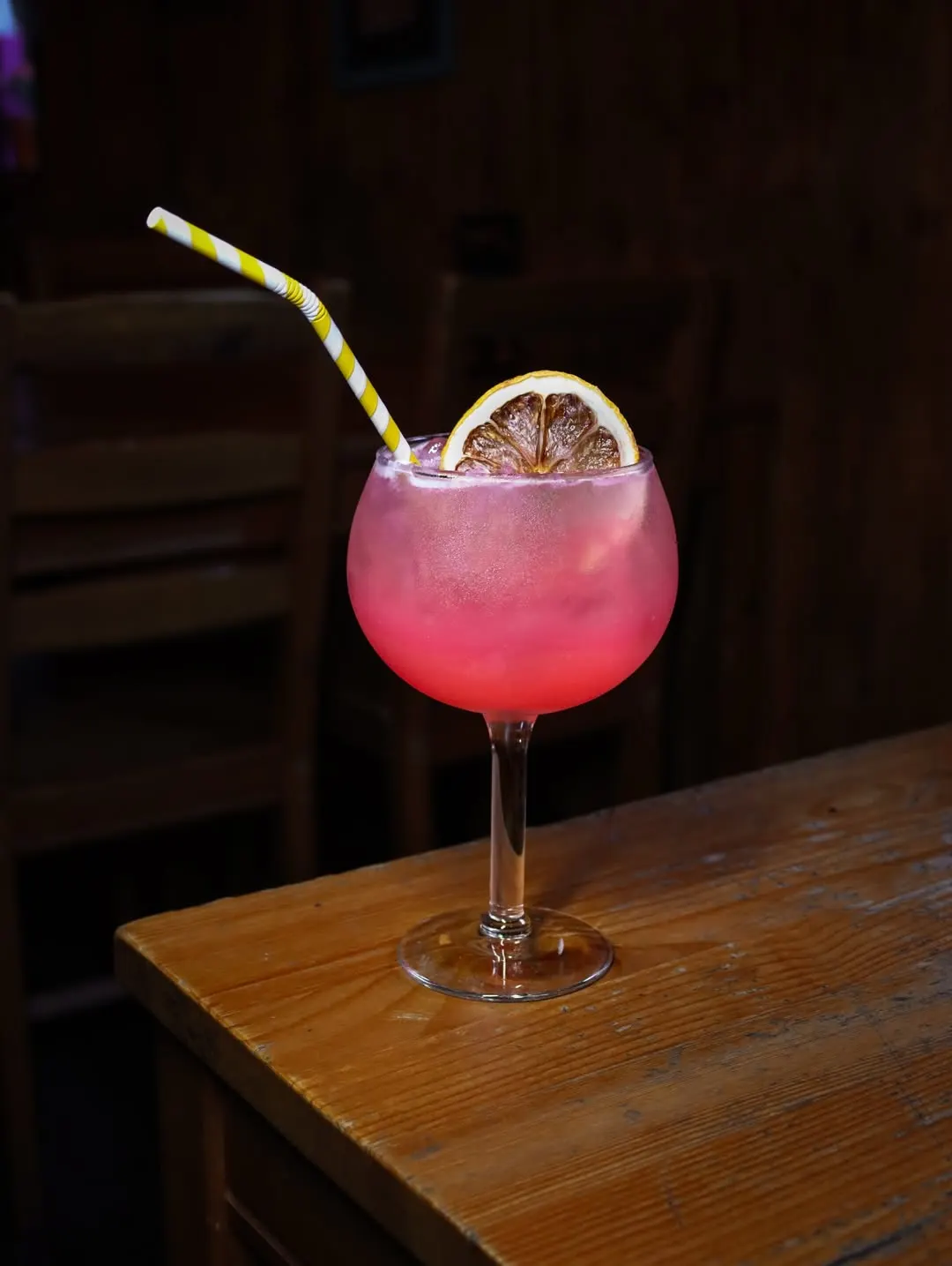 Um copo balão com uma bebida cor-de-rosa, uma rodela de laranja desidratada e uma palhinha às riscas amarelas, sobre uma mesa de madeira.