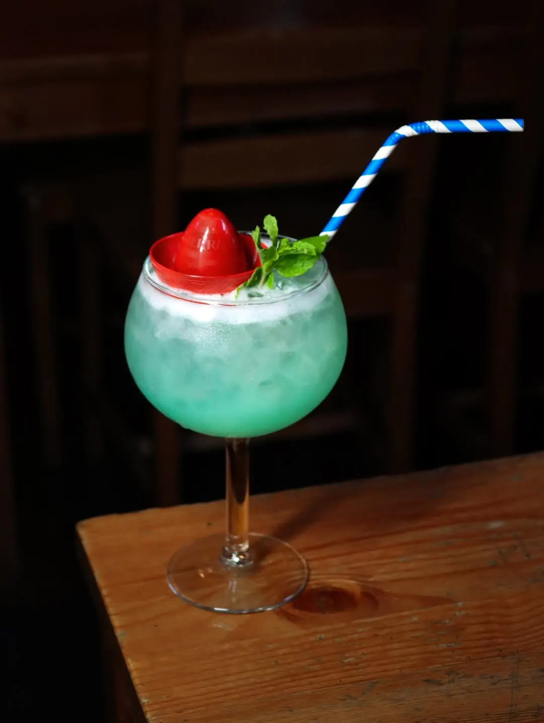 Um copo balão com uma bebida turquesa, gelo picado, um morango, hortelã e uma palhinha às riscas azuis, sobre uma mesa de madeira.