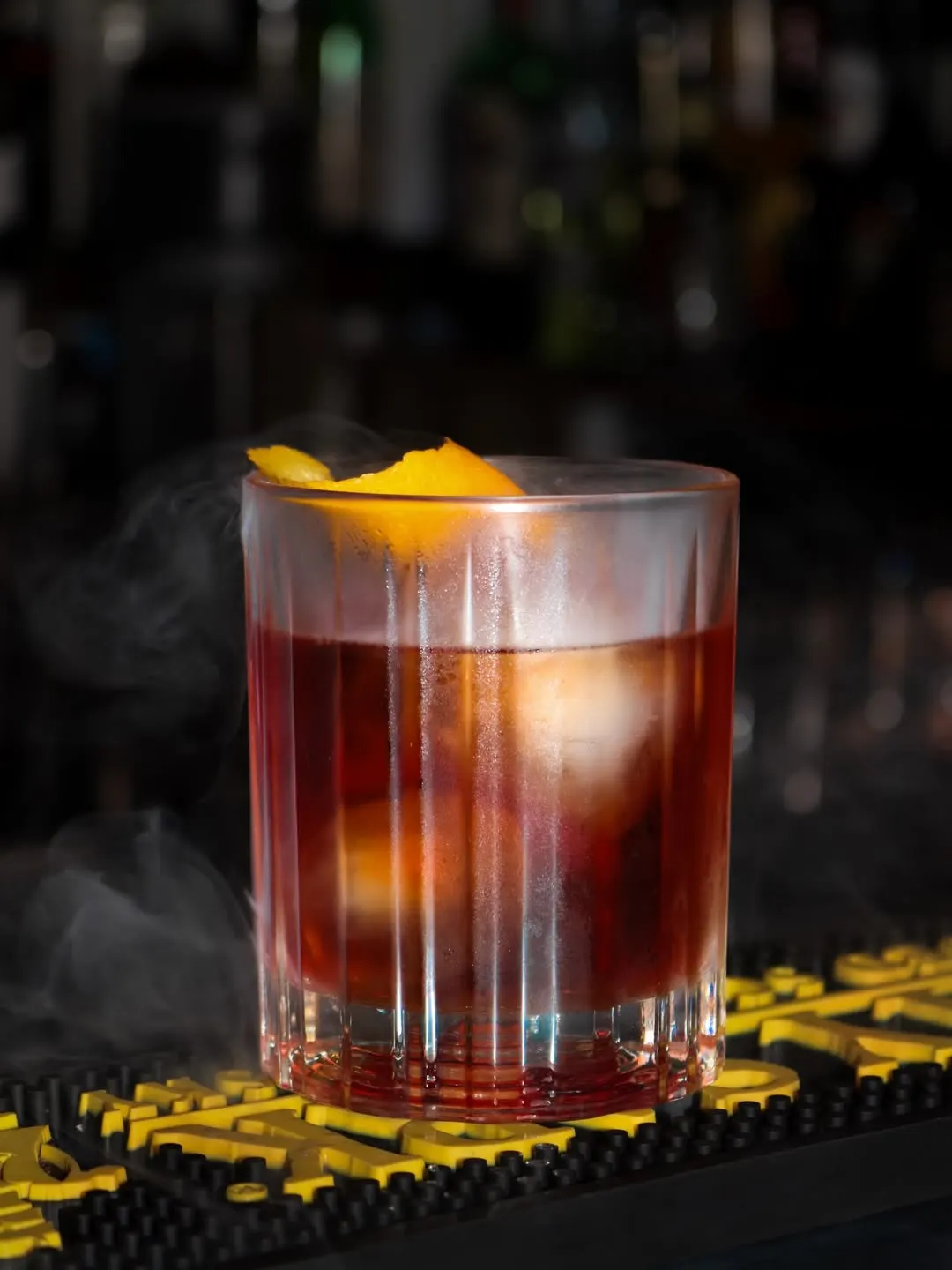 Um copo baixo com uma bebida vermelho-escura, gelo e uma casca de laranja, com fumo à volta, sobre o tapete do balcão.